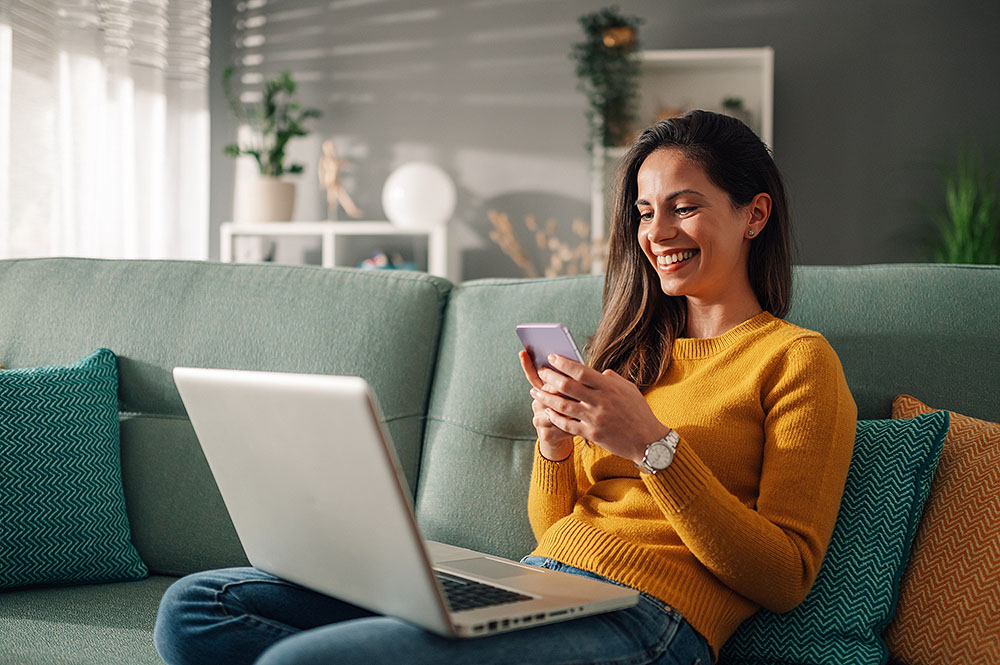 Woman sitting on a sofa at home, using a laptop and phone to research gummy vitamins